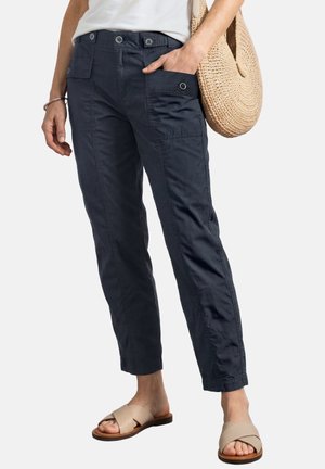 Kvinde iført mørkeblå knap-lukke cargo bukser, beige slip-on sandaler, hvid top og bærer en rund flettet stråtaske på den ene skulder.