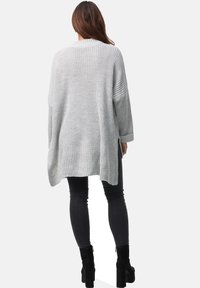 Hellgrauer, übergroßer Strickpullover mit Rundhals, langen Ärmeln, seitlichen Schlitzen und strukturiertem Finish, getragen über schwarzen Jeans und Stiefeletten mit Absatz.