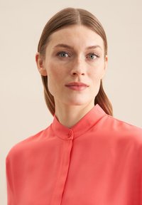 Blusa corallo con bottoni e colletto alla mandarina. Tessuto leggero e liscio con una leggera lucentezza. Design semplice, senza stampati o accenti aggiuntivi.