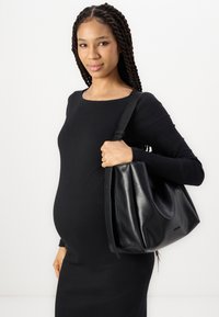 Sac hobo en cuir noir avec une texture douce et une seule sangle, porté au bras, associé à une robe noire ajustée à côtes.