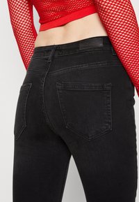 Vaqueros negros ajustados con una textura suave, que cuentan con dos bolsillos traseros y costuras en contraste. La cinturilla muestra una etiqueta de cuero con la marca.