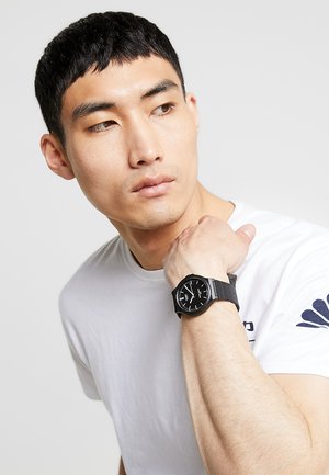 Montre noire avec un cadran rond, rehauts argentés et un bracelet texturé, portée au poignet. T-shirt blanc avec un petit emblème sur la manche.