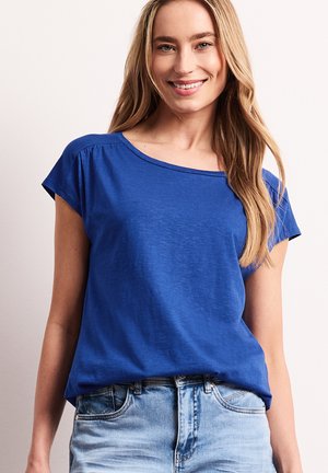 Uśmiechnięta kobieta z długimi blond włosami, ubrana w luźną niebieską koszulkę z krótkim rękawem i jasnoniebieskie dżinsy na jednolitym tle.