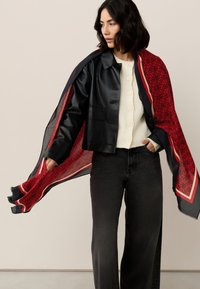 Black faux leather cropped jacket over a cream buttoned sweater, paired with wide-leg black jeans and a red patterned scarf draped over shoulders.