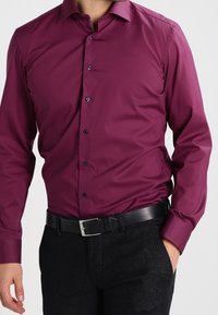 Chemise bordeaux à boutons, avec un col classique, des manches longues et des boutons bleus. Portée avec une ceinture noire et un pantalon texturé foncé.