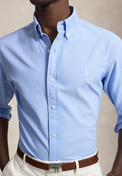 Homme portant une chemise bleu clair à boutons avec manches retroussées, logo blanc brodé sur la poitrine, ceinture en cuir marron et pantalon blanc.