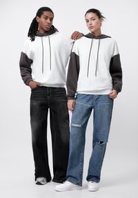Dos modelos llevan sudaderas con bloques de color gris y marrón oscuro, combinadas con vaqueros sueltos negros y azul claro. La sudadera tiene cordones.