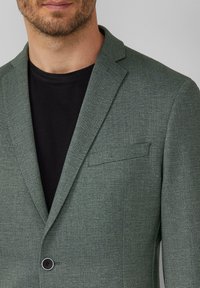 Blazer vert sur mesure en tissu texturé, fermeture à un bouton, et poche poitrine, superposé à une chemise noire.