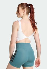Sostén deportivo blanco con un diseño de espalda pronunciada, combinado con pantalones cortos teal de tiro alto con franjas laterales negras. Textura suave, estilo ajustado.