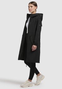 Zwarte lange jas met een capuchon, voorzien van een rits aan de voorkant, zijzakken en elastische manchetten. Draagt over een wit topje en zwarte leggings met witte sneakers.