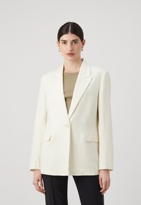 White blazer with a structured design, single button closure, and two front patch pockets; layered over a muted green top.