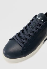 Baskets en cuir bleu marine avec des accents perforés, dotées de lacets plats bleu foncé et d'une semelle en caoutchouc blanc. Le nom de marque "REPLAY" est visible.