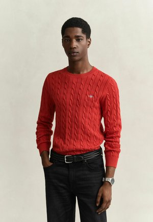 Jeune homme portant un pull à torsades rouge et un pantalon noir, debout avec une main dans la poche devant un fond clair uni.