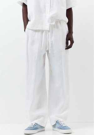 Osoba nosząca luźne białe lniane spodnie i dopasowaną białą koszulę, w połączeniu z niebiesko-białymi casualowymi trampkami, stoi na białym tle.