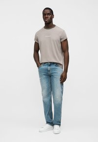 Γκρι κοντομάνικο μπλουζάκι με γυρισμένα μανίκια, συνδυασμένο με ανοιχτό μπλε τζιν παντελόνι με φθορές και λευκά αθλητικά παπούτσια. Το μοντέλο ποζάρει με τα χέρια στις τσέπες.