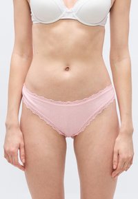 Γυναίκα που φοράει λευκό σουτιέν με δαντέλα και ανοιχτό ροζ εσώρουχο με δαντέλα, στέκεται μπροστά σε ένα απλό φωτεινό φόντο.