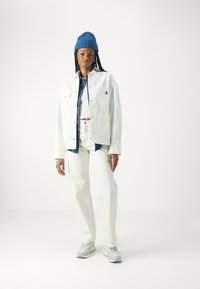 Veste en denim blanche et pantalon, bonnet côtelé bleu et chaussures de sport grises. La tenue comprend une chemise en denim superposée en dessous.