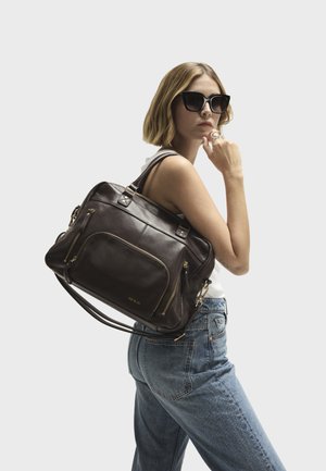 Sac à main en cuir brun foncé avec des accessoires en or, double anse supérieure et plusieurs poches zippées. Modèle portant une tenue décontractée.