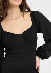 Femme portant une robe noire texturée avec des manches longues bouffantes et un décolleté carré froncé, main reposant sur la hanche.