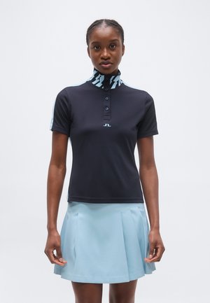 Femeie purtând un polo negru cu mânecă scurtă și guler cu model, și o fustă plisată albastru deschis, stând în picioare pe un fundal alb simplu.