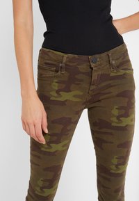 Kamouflagejeans i nyanser av grönt och brunt, med slim fit, framfickor och metallknappstängning. Mjuk, texturerad tyg.