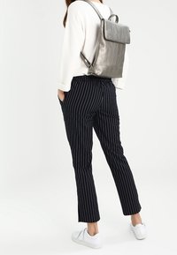 Henkilö, joka on pukeutunut tummansinisiin raidallisiin housuihin, valkoisiin lenkkareihin, valkoiseen pitkähihaiseen puseroon ja metallinhohtoiseen hopeiseen reppuun, seisoo kädet taskuissa.