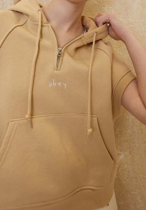 Sudadera beige de manga corta con cremallera frontal y bolsillo canguro, texto bordado "obey", persona sujetando la capucha cerca del cuello.