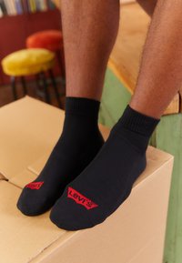 Schwarze Baumwollsocken mit gerippten Bündchen und einem roten "Levi's"-Logo auf der Oberseite. Getragen mit nackten Füßen, die auf einem Karton stehen.