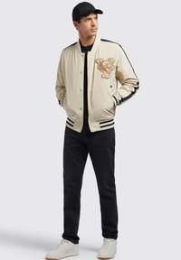 Beige bomberjakke med et tigerpatch, trykknapper, sorte striber på ærmerne og ribbede manchetter. Bæret med sorte jeans og hvide sneakers.