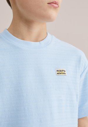 Jaunas žmogus dėvi šviesiai mėlyną tekstūruotą marškinėlį su mažu "PUERTO MONTAÑA" lipduku kairėje krūtinės pusėje.