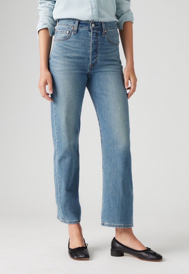 Personne portant un jean droit bleu clair, une chemise bleu pâle avec les manches retroussées, et des ballerines noires à petit talon.