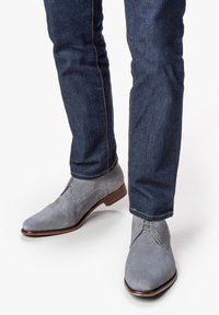 Graue Stoffschuhe mit glatter Textur zeichnen sich durch ein gesticktes Design und Schnürverschluss aus, ergänzt durch eine braune Ledersohle und -akzente.