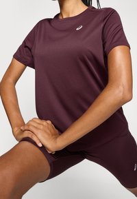 Person som har på sig en maroon träningsskjorta och shorts, sträcker ut benen med händerna knäppta på låret, mot en helt vit bakgrund.