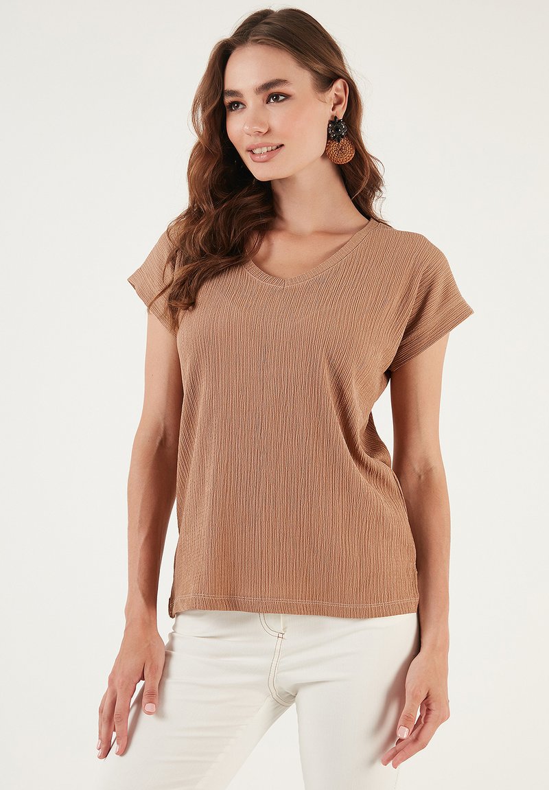 Brown, short-sleeve top with a V-neck in textured fabric, featuring subtle vertical stripes. Worn with white pants, showcasing relaxed fit.