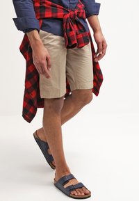 Homme portant un short beige, des sandales bleu marine, une chemise bleu marine et une chemise à carreaux rouge et noir nouée autour de la taille, se tenant sur un fond blanc.