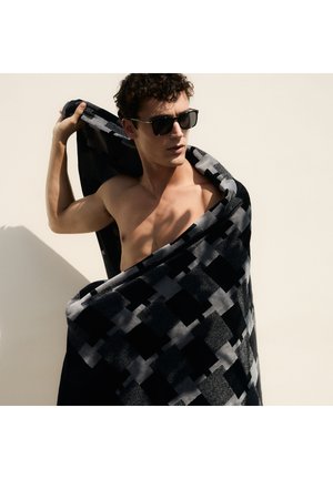 Homme torse nu portant des lunettes de soleil, enroule une grande serviette à carreaux noirs et gris autour de ses épaules, sur un fond clair uni.