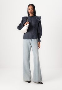 Μπλε σκούρη μπλούζα με βολάν στα μανίκια και υφή στο μπροστινό μέρος, συνδυασμένη με ανοιχτό γαλάζιο καμπάνα τζιν και λευκή τσάντα. Μαύρα μυτερά παπούτσια ολοκληρώνουν το σύνολο.