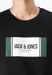 Svart bomull T-skjorte med et sentralt hvitt rektangulært logo med grønne detaljer og teksten "JACK & JONES COLLECTIONS EST. 1990."