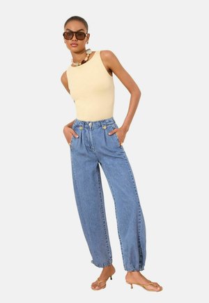 Nainen, joka pitää päällään hihattoman beigen topin, korkeavyötäröiset, väljät siniset farkut nappidetaljeilla, ruskeat korolliset sandaalit, isot aurinkolasit ja simpukkakaulakorun.