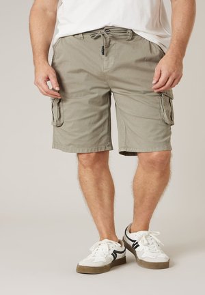 Homme portant un short cargo marron clair avec poches latérales, des baskets blanches avec des détails noirs, et une chemise blanche, debout devant un fond uni.