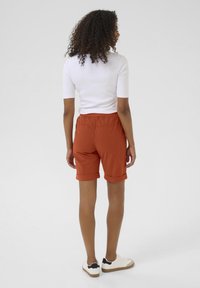 Shorts couleur rouille avec ourlets roulés, taille élastique et tissu léger, associés à un haut ajusté blanc et des baskets.