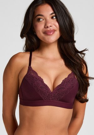 Vrouw met lang donker haar die een bordeauxrode kanten bralette draagt, glimlacht en kijkt iets naar links tegen een effen achtergrond.