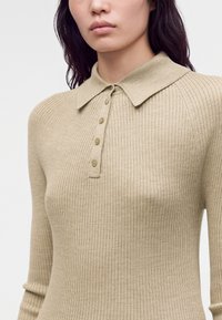 Femme portant un top beige en maille côtelée à manches longues avec un col et quatre boutons sur la patte de boutonnage avant.