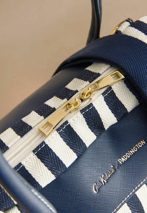 Tuvplāns uz tumši zilu un baltu svītru somu ar zelta dubulto rāvējslēdzēju un "Cath Kidston PADDINGTON" logotipu uz teksturētas virsmas.