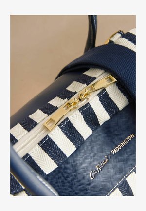 Közeli kép tengerészkék-fehér csíkos táskáról, arany dupla cipzárral és a "Cath Kidston PADDINGTON" logóval, texturált felületen.