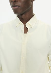 Chemise jaune pâle à boutons avec une texture douce, présentant un col classique et une fermeture à un seul bouton, montrée sur un fond uni.