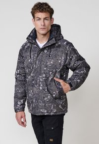 Chaqueta acolchada negra y gris con un patrón, capucha, bolsillos frontales y cordones ajustables. El material presenta una superficie texturizada.