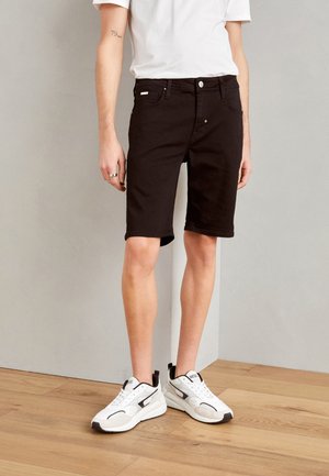 Pantalones cortos de mezclilla negra con corte recto, que cuentan con un cierre de botón frontal, bolsillos laterales y un dobladillo que termina justo por encima de la rodilla. Combinados con zapatillas blancas.