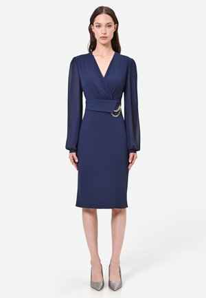 Femme portant une robe bleu marine longueur genou avec manches longues transparentes et un devant style cache-cœur avec détail chaîne dorée à la taille.