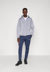 Ljusgrå zip-up jacka med huva, matchad med marinblå sweatpants med en vit logotyp. Svarta sportskor fulländar outfiten.